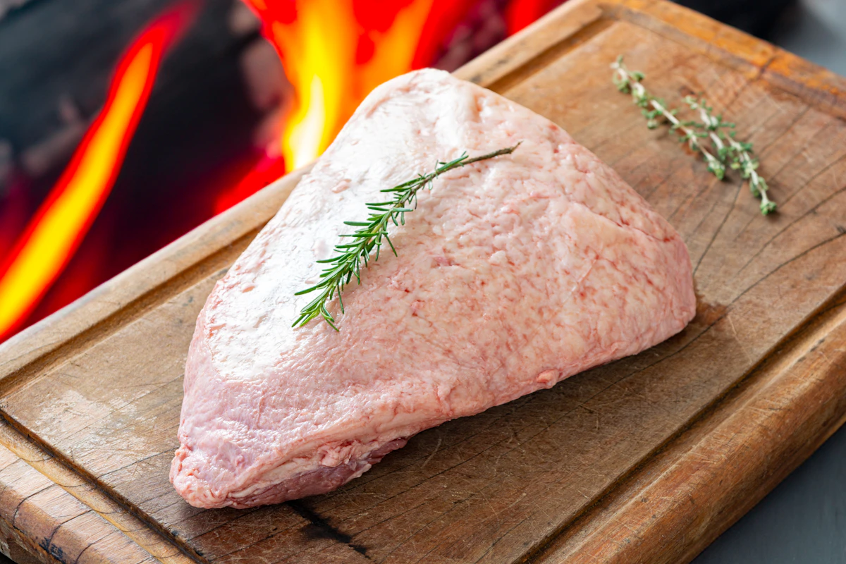 Peça de picanha brasileira crua sobre tábua de madeira com a capa de gordura aparente e ramo de alecrim ao lado, preparada para o churrasco